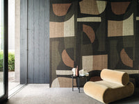 Woonkamer met Concordance behang van Casamance in een groot abstract geometrisch patroon met zwart, kaki en aardetinten, gecombineerd met een lichte loungestoel.