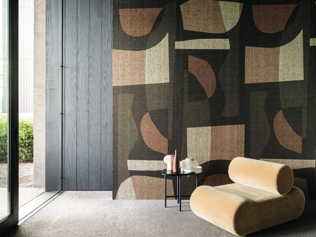 Woonkamer met Concordance behang van Casamance in een groot abstract geometrisch patroon met zwart, kaki en aardetinten, gecombineerd met een lichte loungestoel.